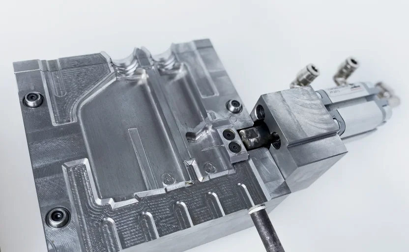 A close-up view of a precision-machined metal mold with intricate cavities and channels, featuring attached pneumatic or hydraulic components, set against a plain white background.