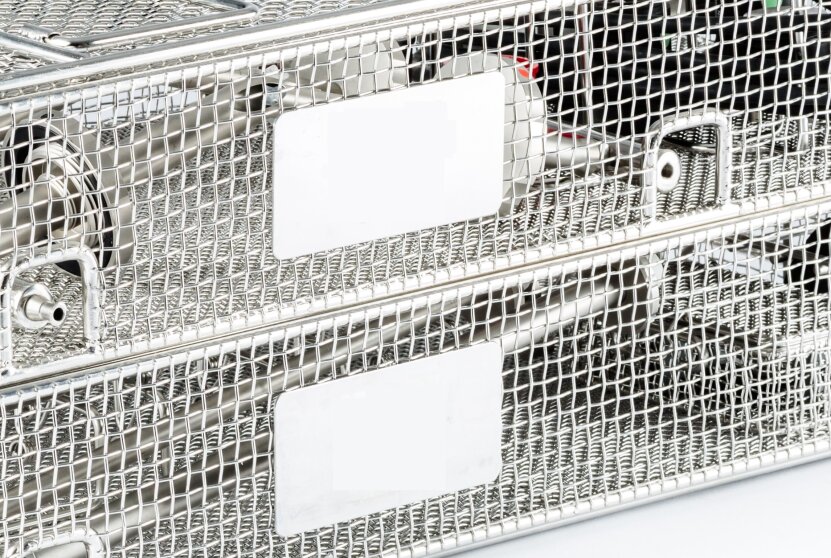 Two stainless steel mesh baskets, stacked one above the other, with blank white labels on the front. The baskets are likely used for storing or sterilizing medical instruments.
