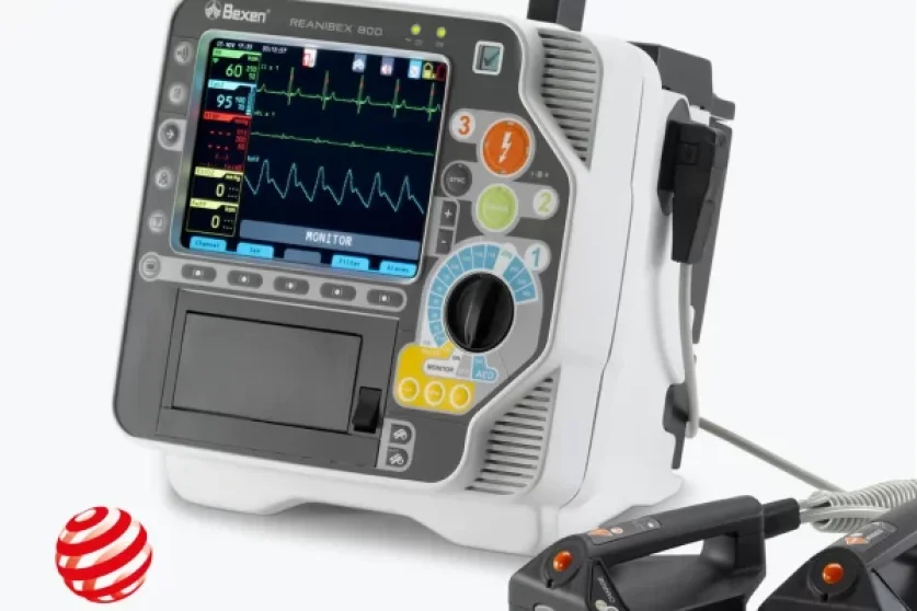 A medical defibrillator monitor displaying vital signs and heart rate, with controls and buttons on the front panel. A handheld defibrillator paddle is attached, and a Red Dot Design Award logo is visible in the bottom left corner.