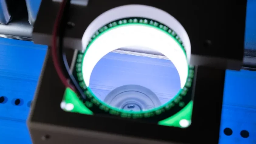 Close-up of a circular machine part with a glowing green light ring, wires attached, and a blue surface beneath, suggesting a high-tech or industrial device in operation.