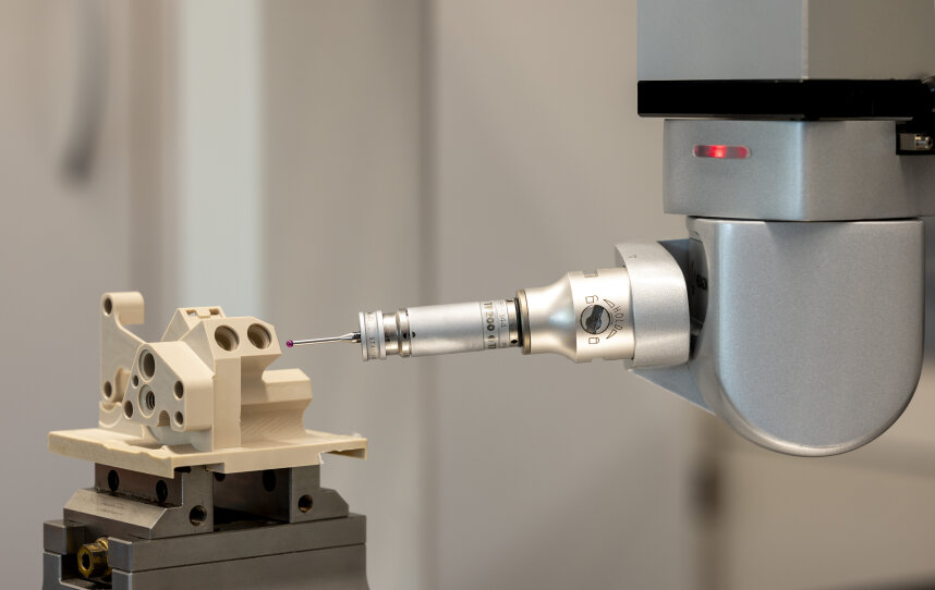 Ein Roboterarm mit einer Präzisionssonde prüft ein beigefarbenes mechanisches Teil, das auf einer Plattform montiert ist, wahrscheinlich für die Qualitätskontrolle in einer Industrie- oder Fertigungsumgebung.
