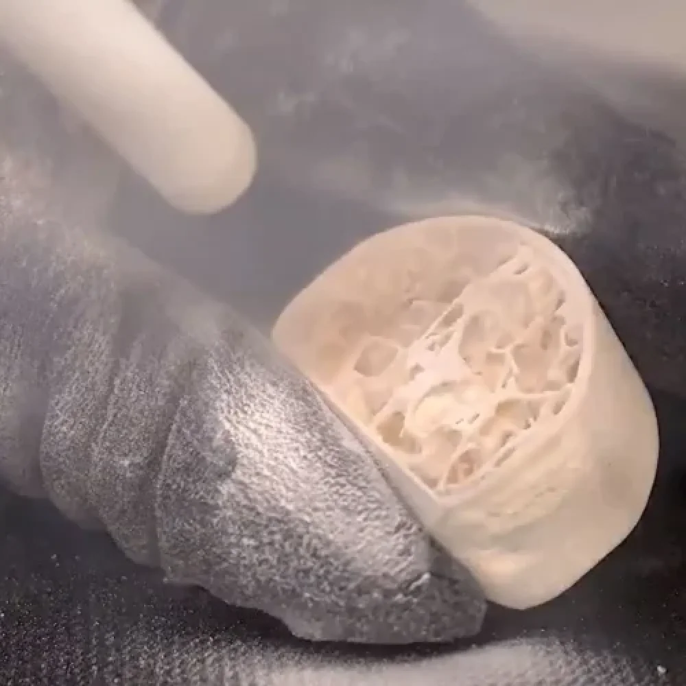 A gloved hand holds a round, cross-sectioned bone while a tool smooths its surface, producing a cloud of fine white dust.