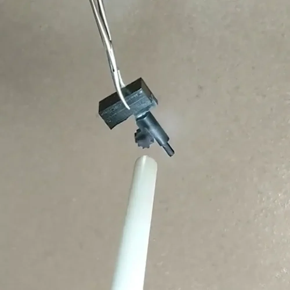 Close-up of tweezers holding a small black plastic gear above the tip of a white cylindrical object, against a plain beige background.