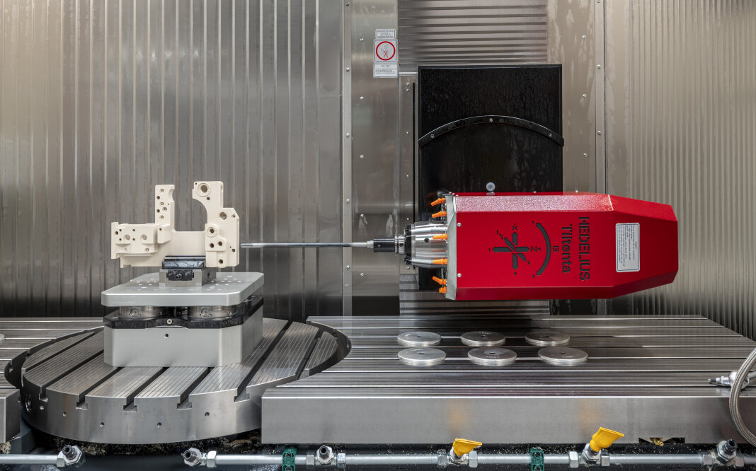 Eine CNC-Maschine in Betrieb mit einem roten Spindelkopf, der sich horizontal in Richtung eines montierten Metallwerkstücks auf einer kreisförmigen Plattform innerhalb eines Metallgehäuses dreht. Scheiben und Maschinenteile sind auf dem Tisch sichtbar.