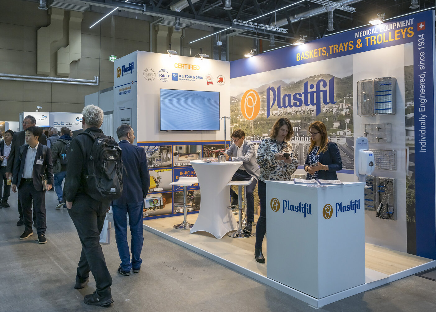 Menschen gehen an einem Messestand von Plastifil vorbei, an dem medizinische Geräte ausgestellt sind. Hinter der Theke stehen zwei Frauen, von denen eine schreibt und die andere spricht; im Hintergrund sind Banner, Tabletts und Produktpräsentationen zu sehen.