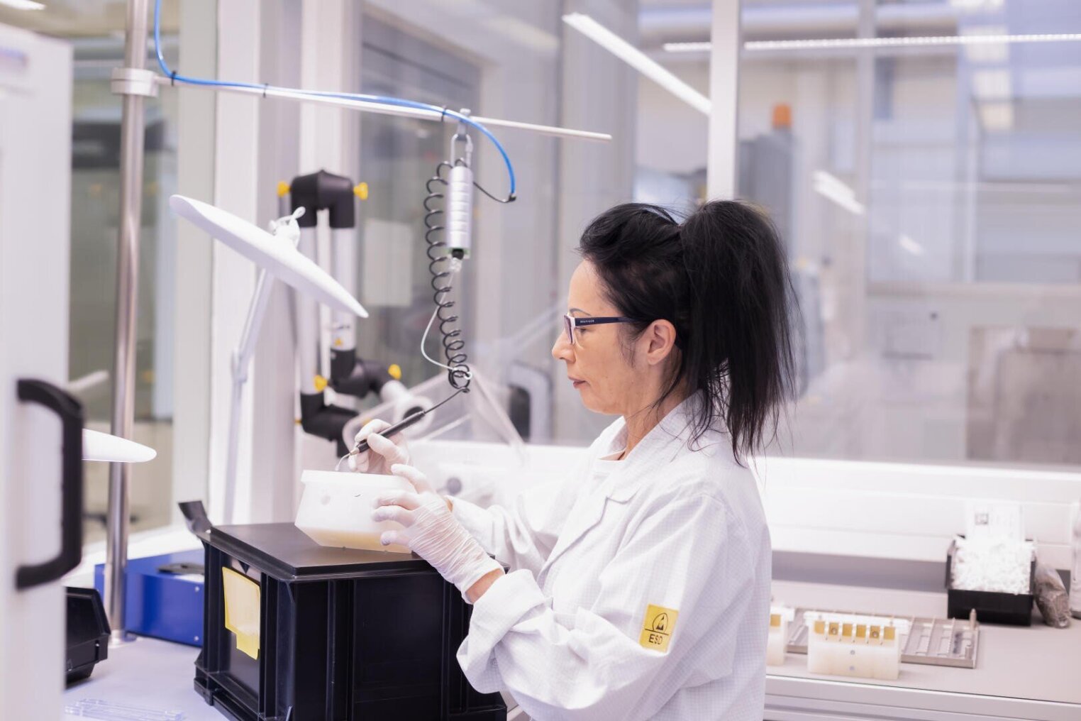 A scientist in a white lab coat and gloves is working with laboratory equipment in a bright, modern laboratory. She appears to be concentrating as she operates a pipette or similar tool near a plastic container.