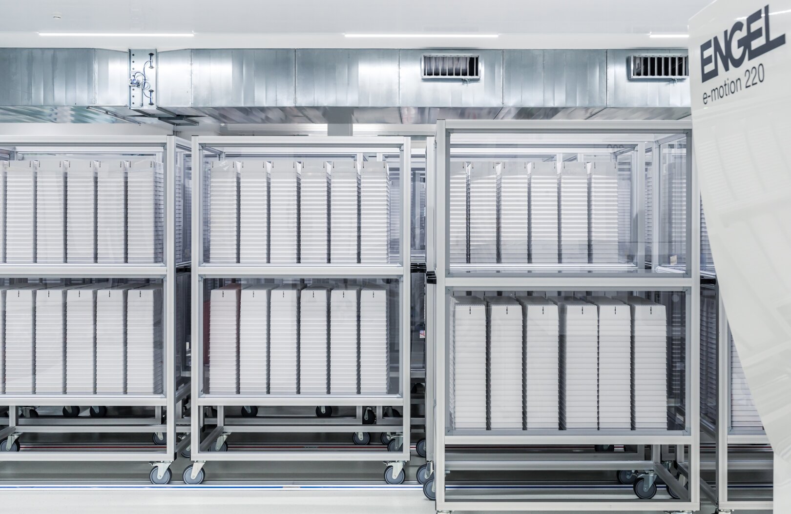 In einer hellen, sauberen Industrieanlage sind Metallregale mit mehreren weißen vertikalen Platten angeordnet. Die Umgebung wirkt modern und steril, und rechts ist teilweise eine Maschine mit der Aufschrift ENGEL e-motion 220 zu sehen.