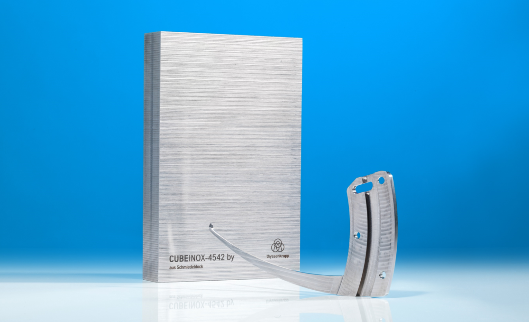 Ein rechteckiger, gebürsteter Metallblock mit der Aufschrift CUBEINOX-4542 steht aufrecht neben einem gebogenen, perforierten Metallstreifen, beide vor einem hellblauen Hintergrund.