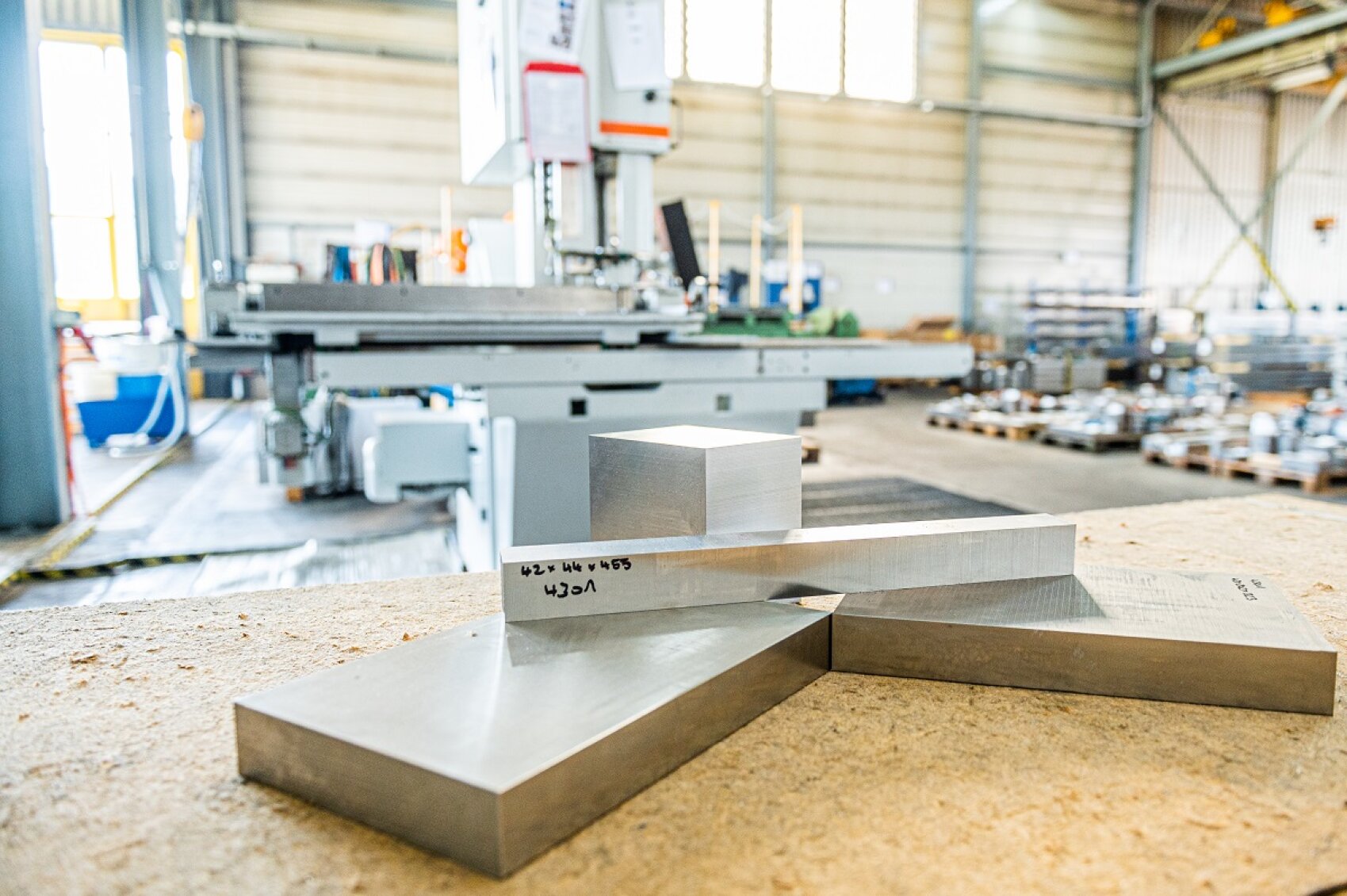 Drei rechteckige Metallblöcke liegen auf einer Werkbank im Vordergrund einer hellen, geräumigen Industriehalle voller Maschinen und Geräte.