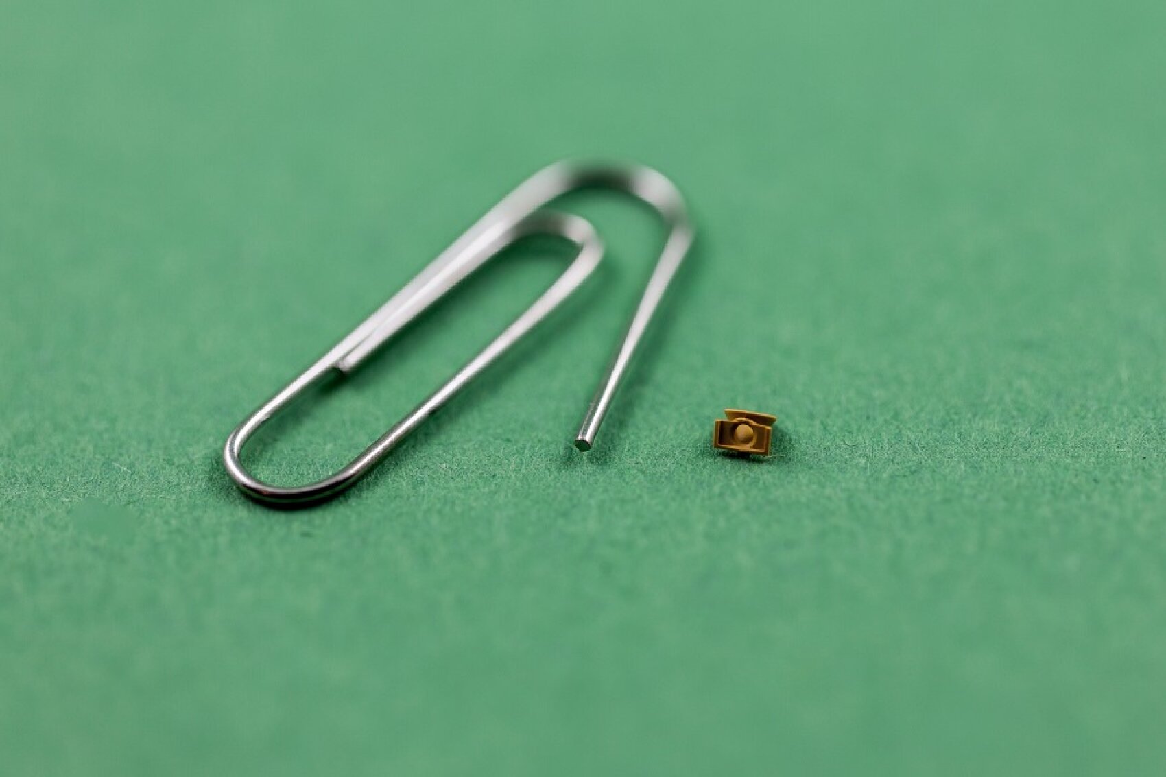A tiny electronic component is placed next to a standard metal paper clip on a green background, emphasizing the small size of the component.