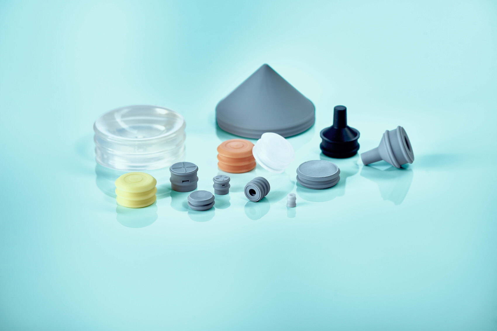A collection of differently shaped and colored rubber and plastic stoppers and closures are arranged on a smooth, light blue surface with a gentle color gradient.