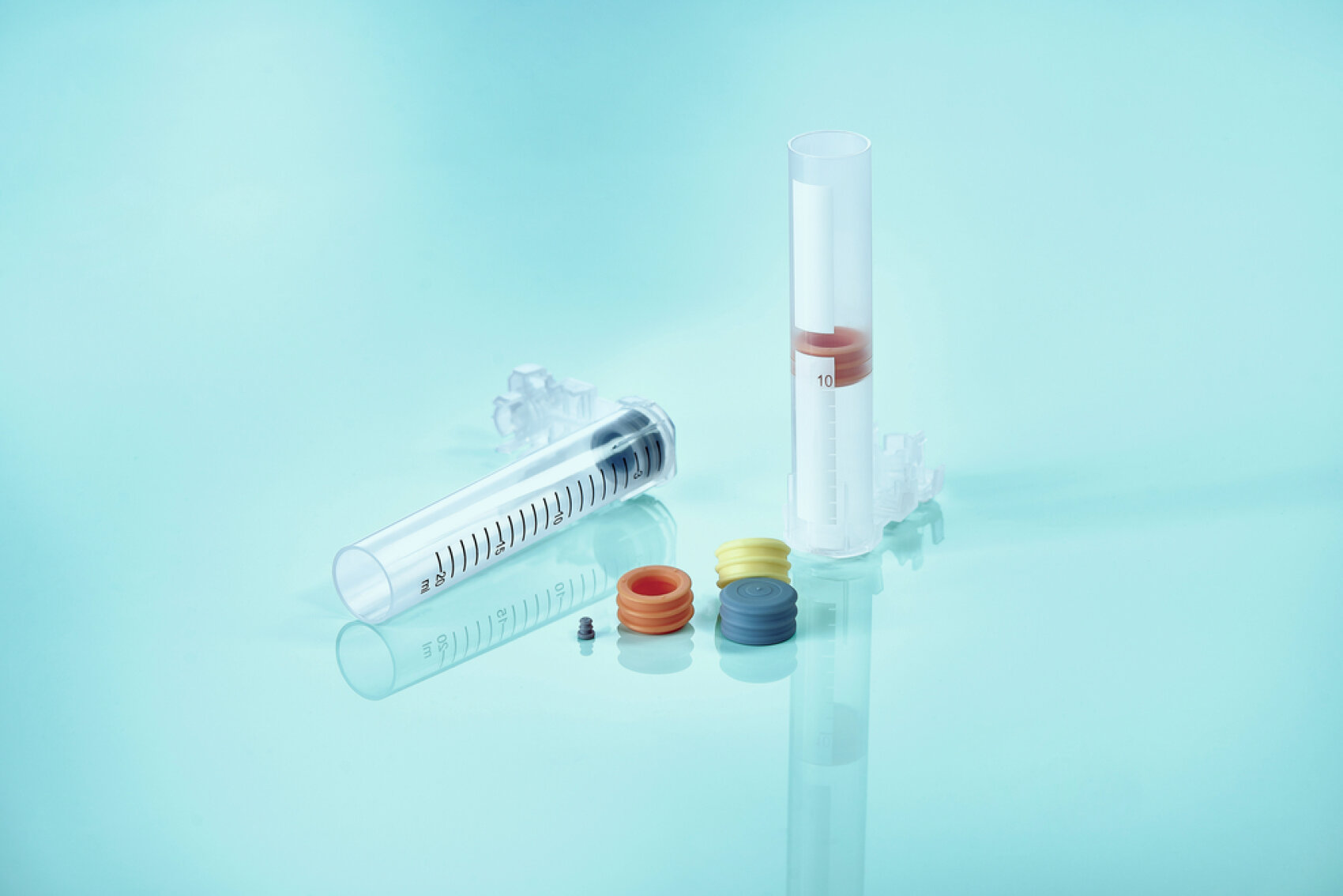 A clear plastic syringe and a transparent cylindrical container are shown alongside several colorful rubber stoppers on a light blue reflective surface.